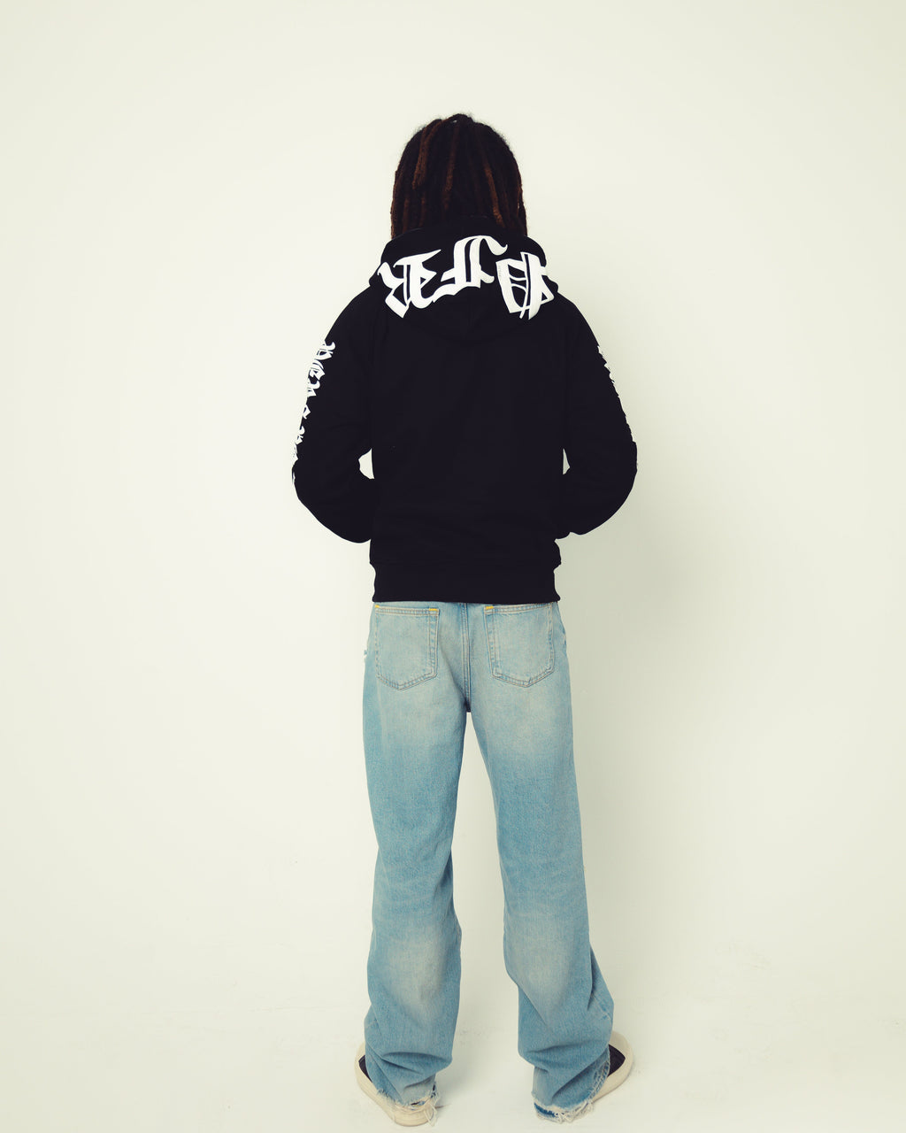 Person wearing a black hoodie with white text and light blue jeans on a white background