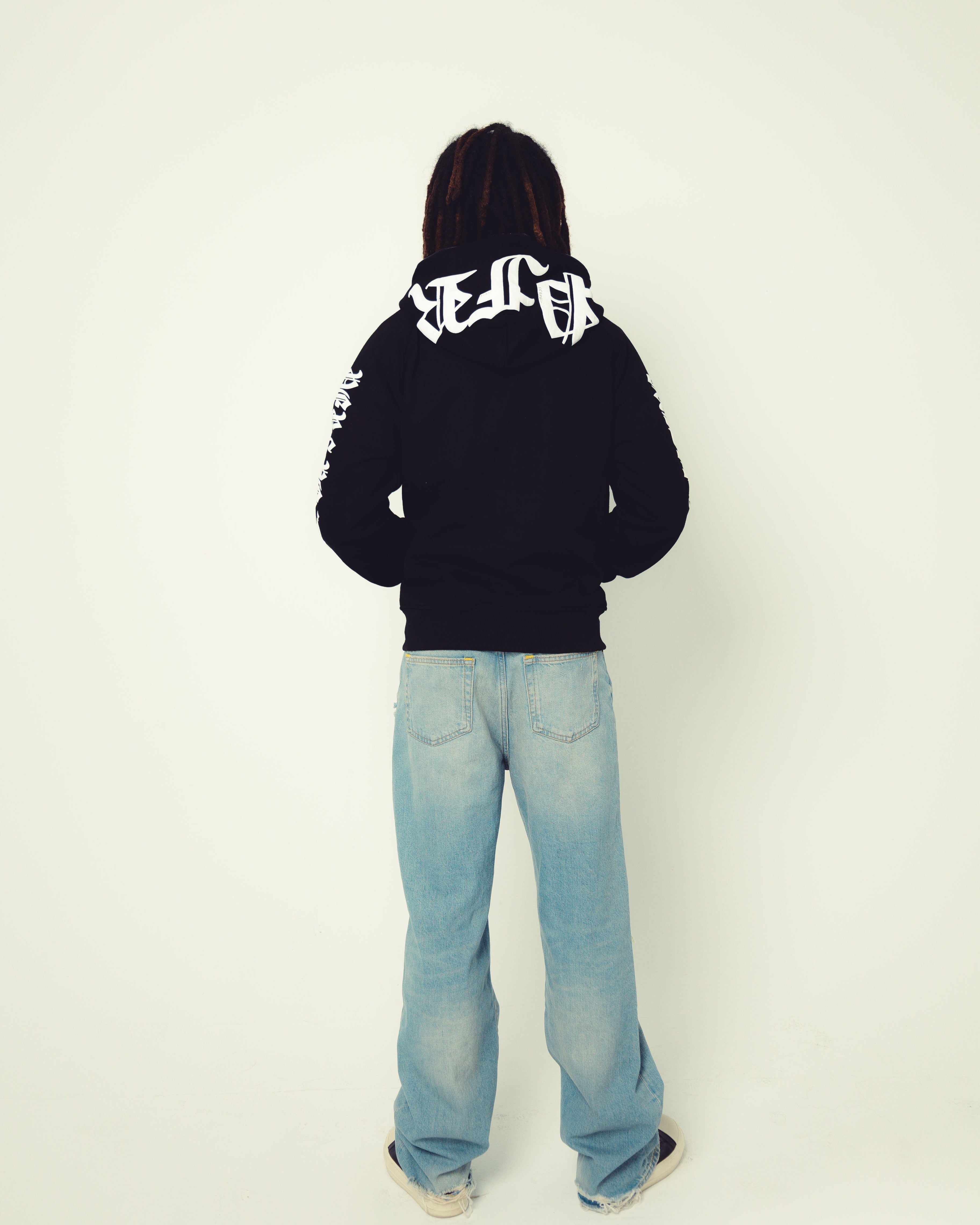 Person wearing a black hoodie with white text and light blue jeans on a white background
