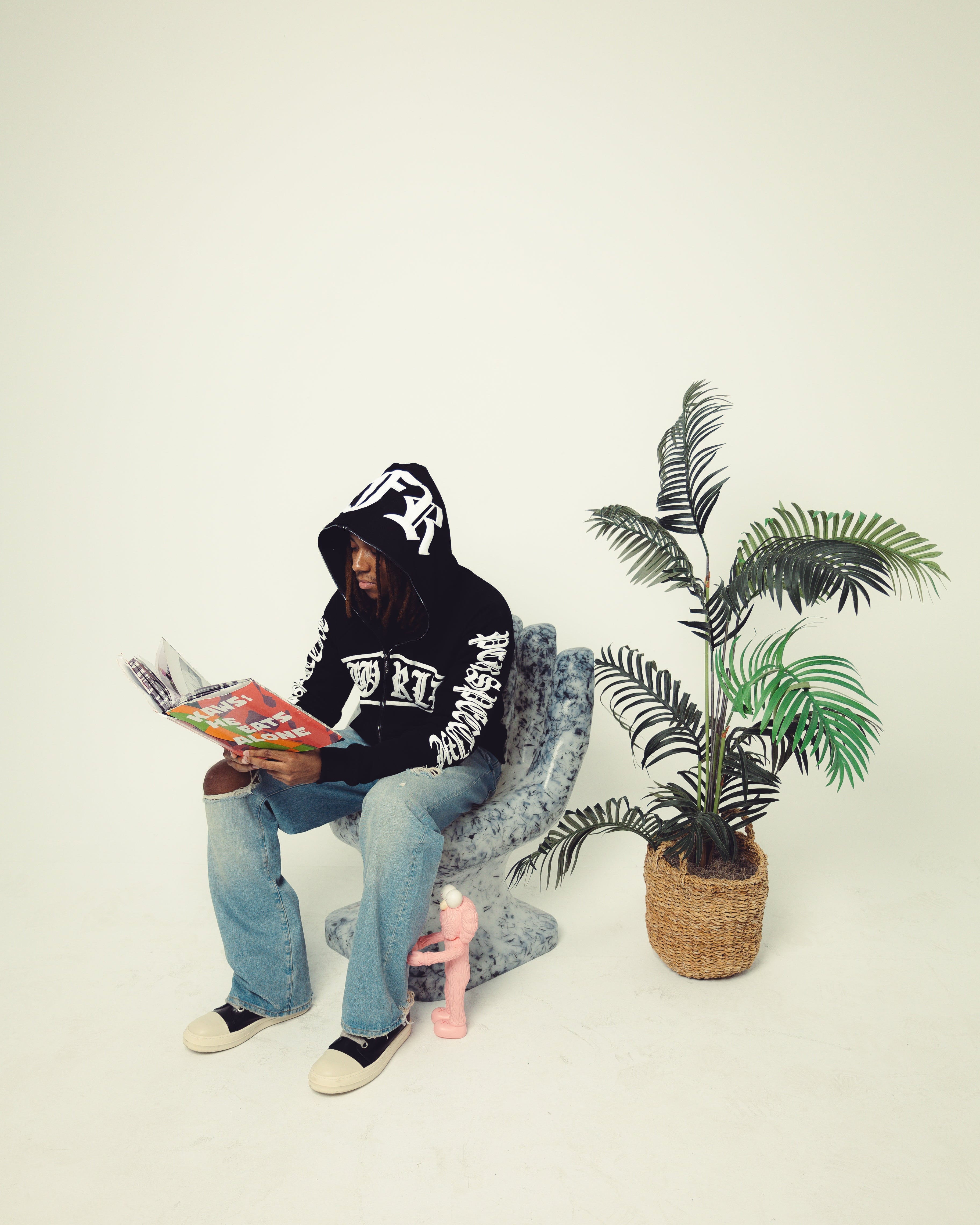 Person sitting on a chair reading a magazine with a plant and pink object in the background