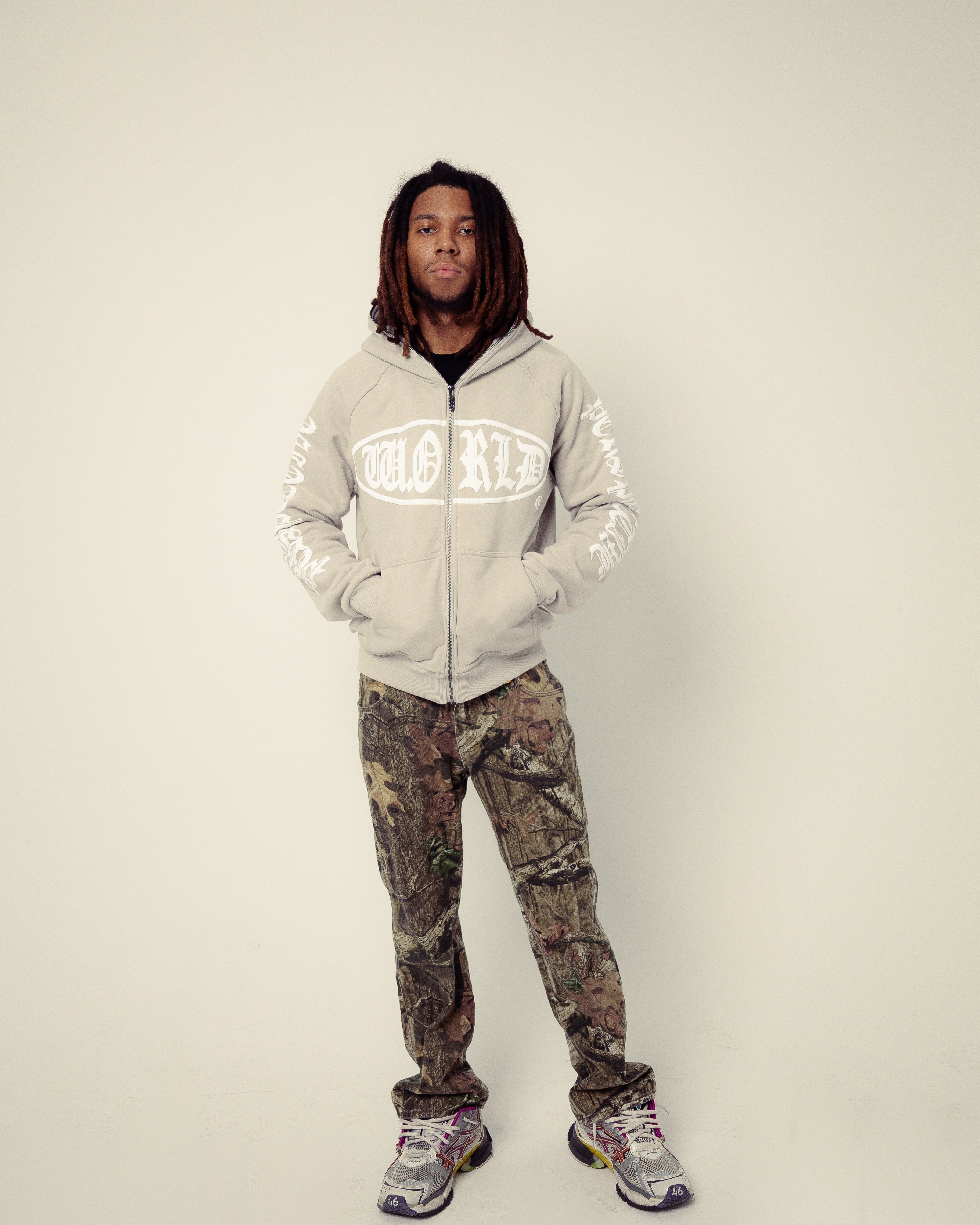 Person wearing a light gray hoodie with text and camouflage pants on a plain background