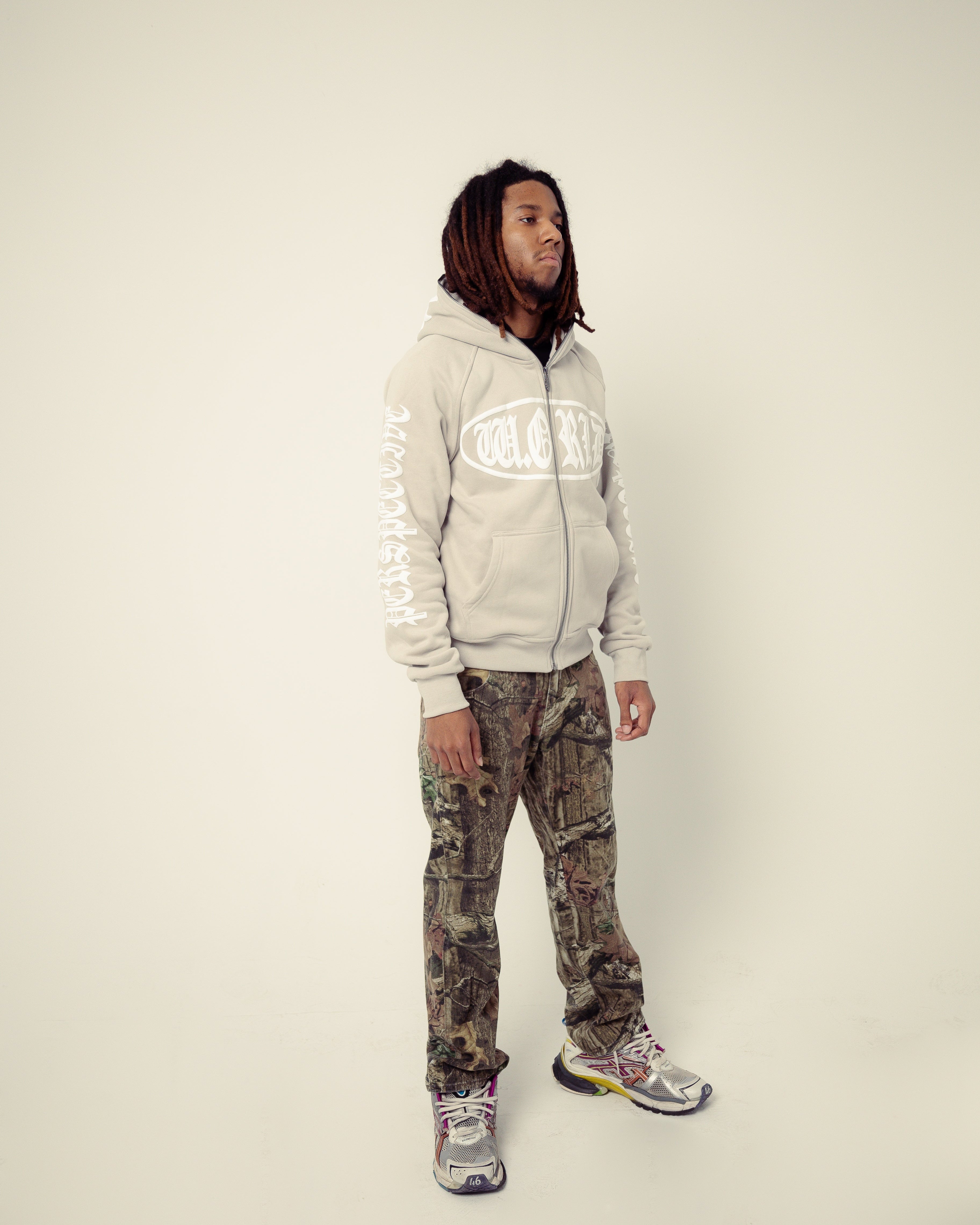 Person wearing a light gray hoodie with a logo and camouflage pants on a plain background