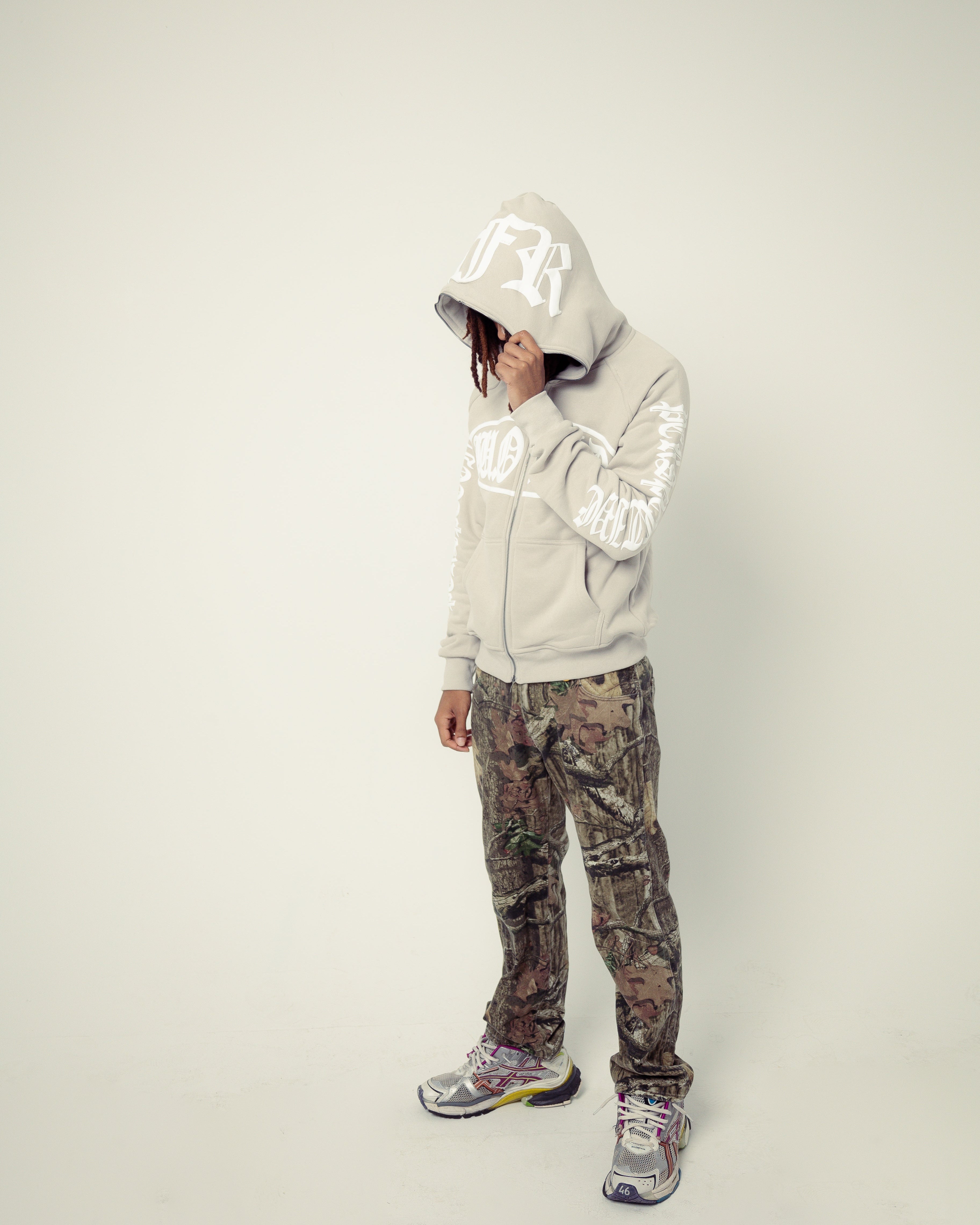 Person wearing a light gray hoodie with a hood and camouflage pants on a white background