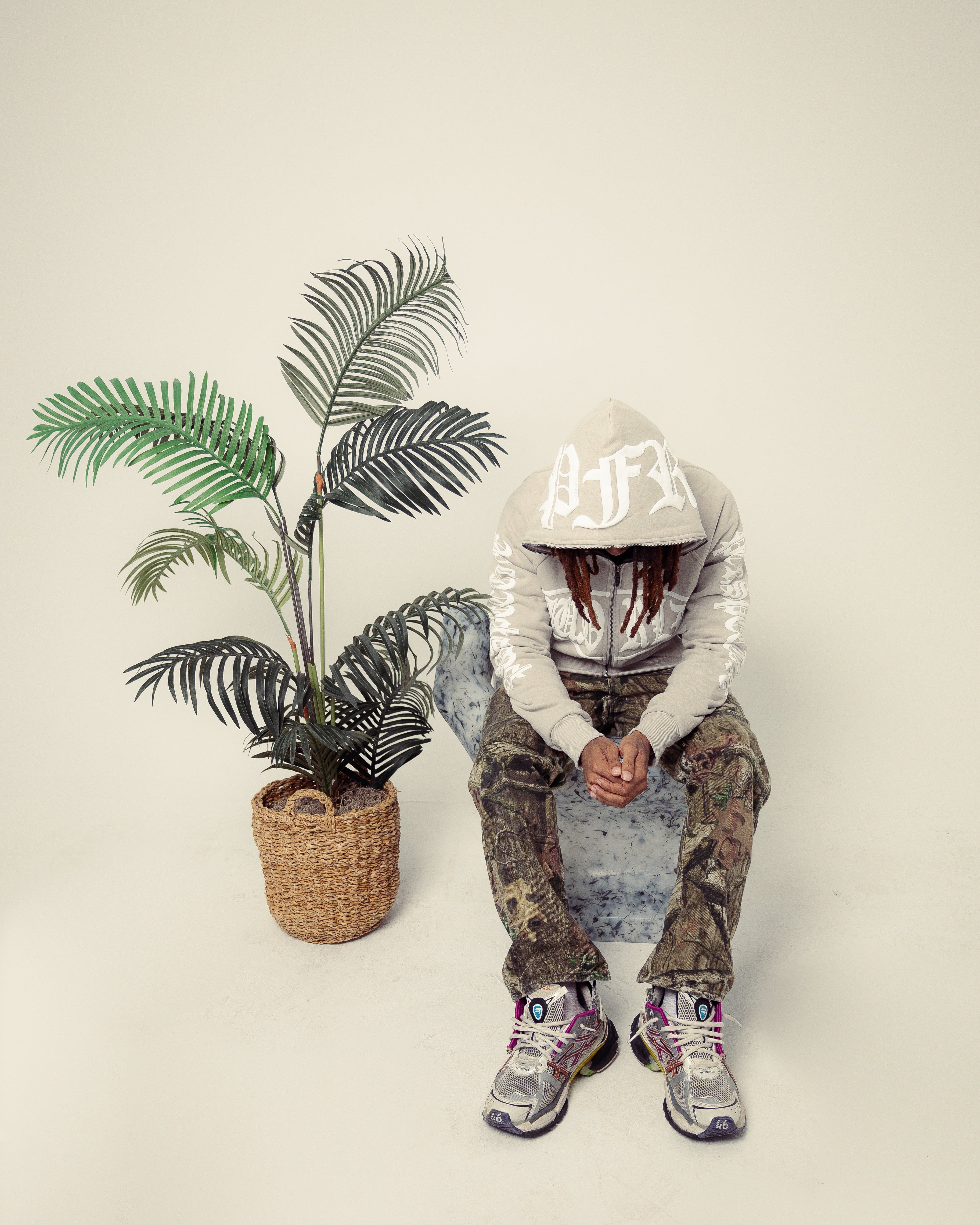 Person wearing a hoodie and camouflage pants sitting next to a potted plant on a plain background