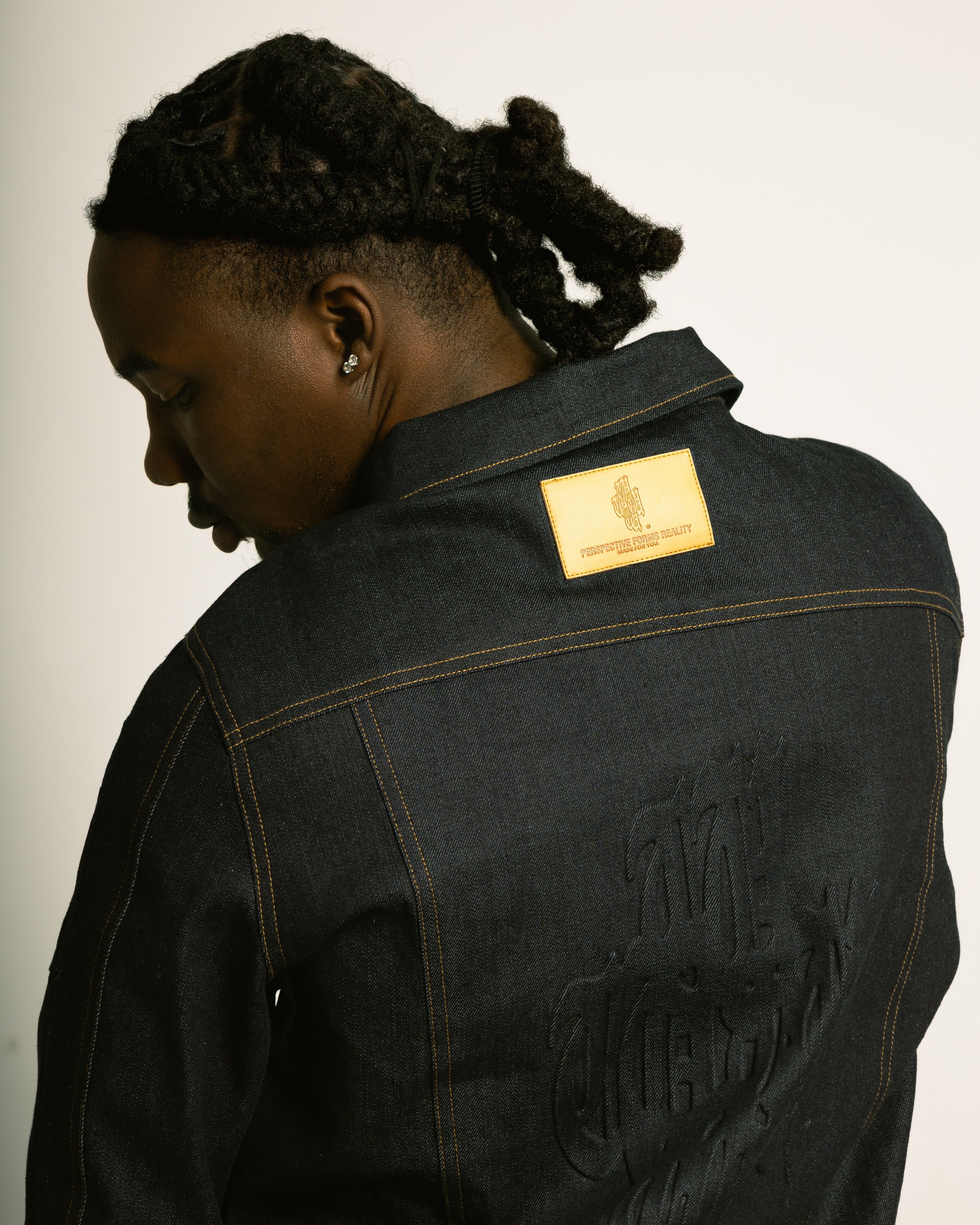 Person wearing a black denim jacket with a gold patch on a plain background
